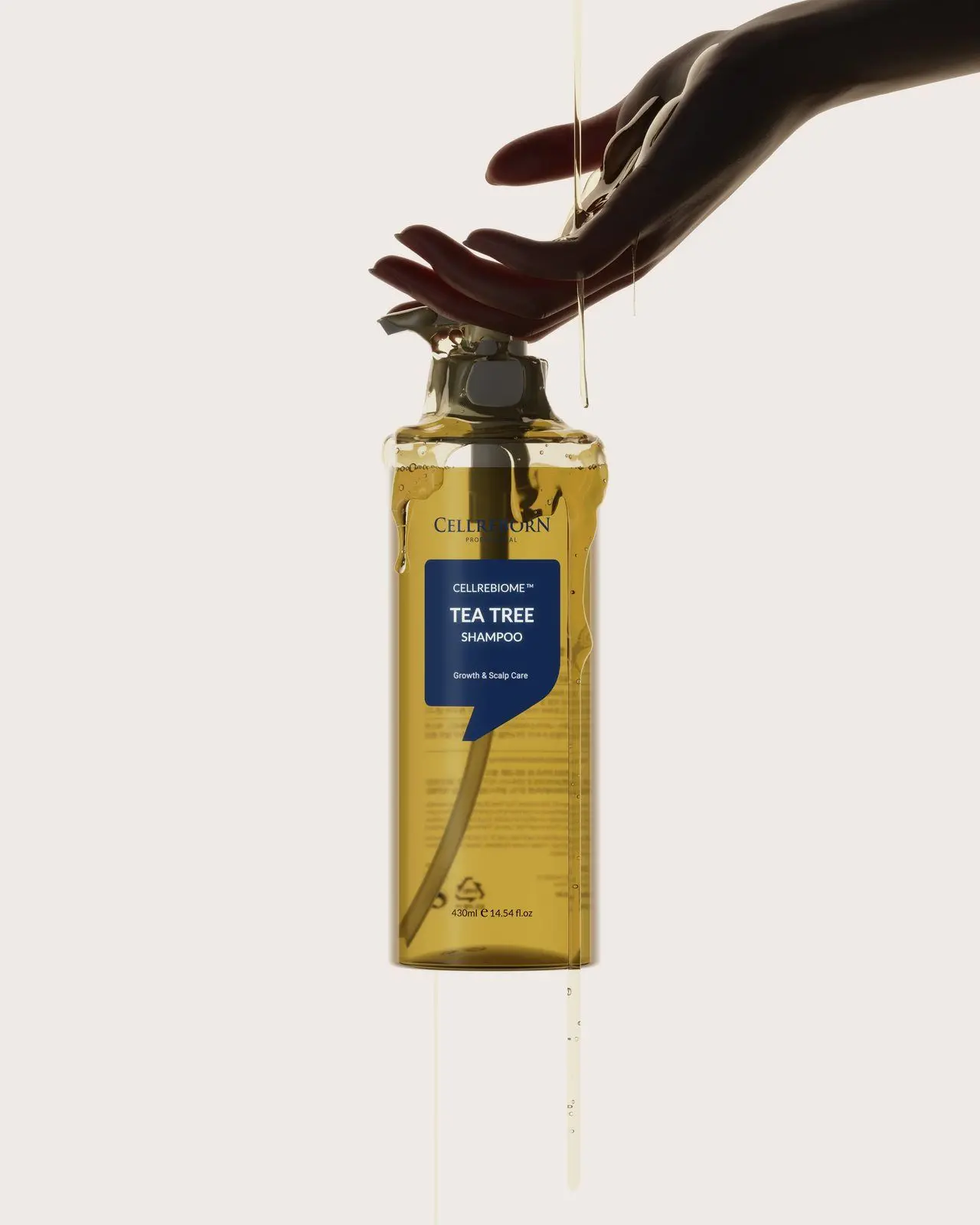 A hand pressing the pump of a Cellrebiome Tea Tree Shampoo bottle with liquid running down, on a cream backdrop